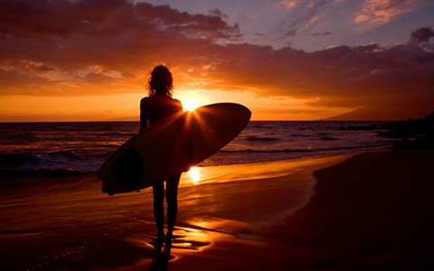 A person standing on a beach at sunrise holding a surfboard